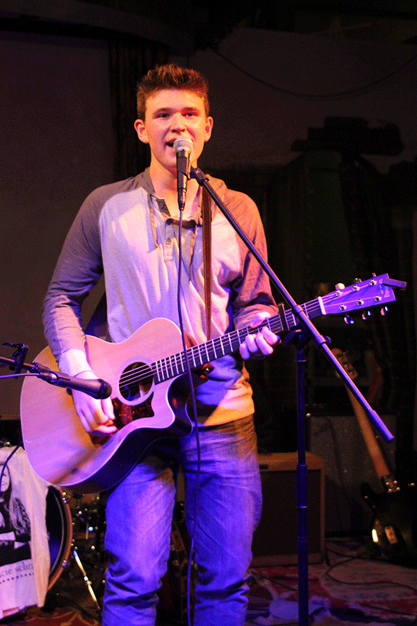 Walsh looks toward the audience as he strums an acoustic guitar and sings.