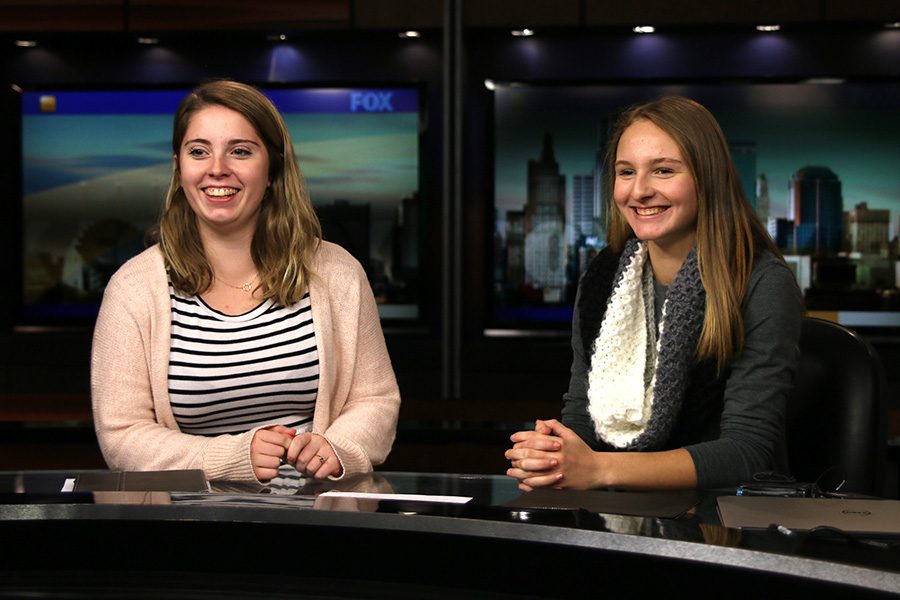 Senior MVTV producers Emma Barge and Bailey Heffernon smile while sitting behind the news desk used by Fox 4 reporters.