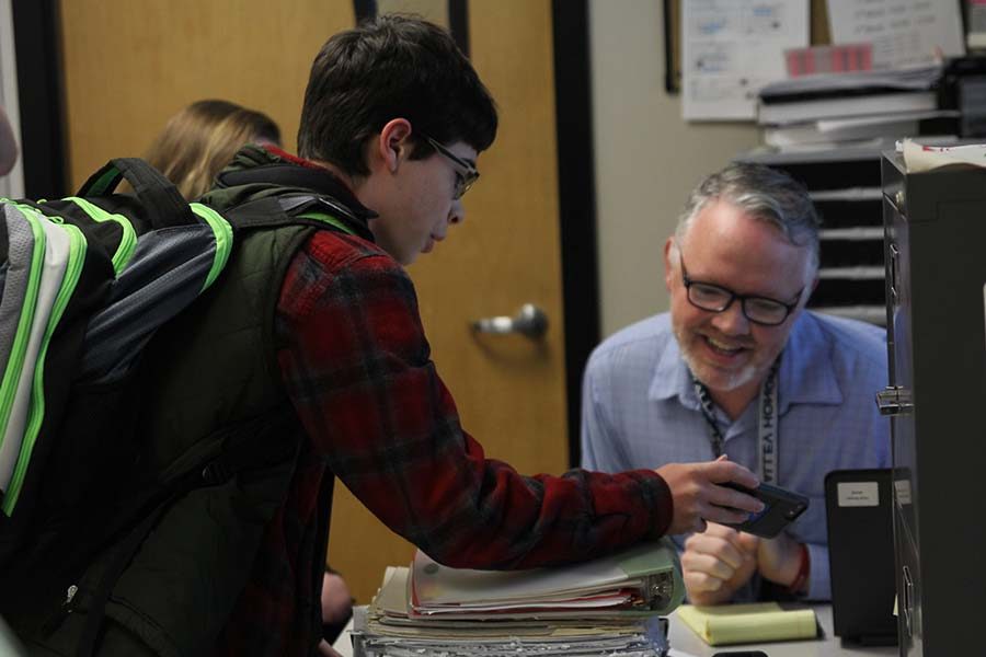 As he leans over the desk, freshman Alex Whipple shows science teacher Chad Brown a video on his phone before school on Tuesday, April 10.