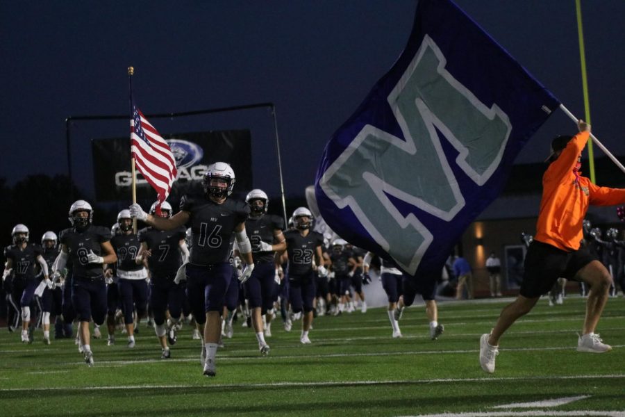 To start the game, defensive back Trey Worcester leads the group of football players to the sideline with the American flag in his right hand.