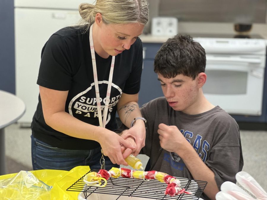 With a smile on her face, paraprofessional Kaila Masters helps senior Jackson Stutheit tie dye a shirt using yellow and red dye.