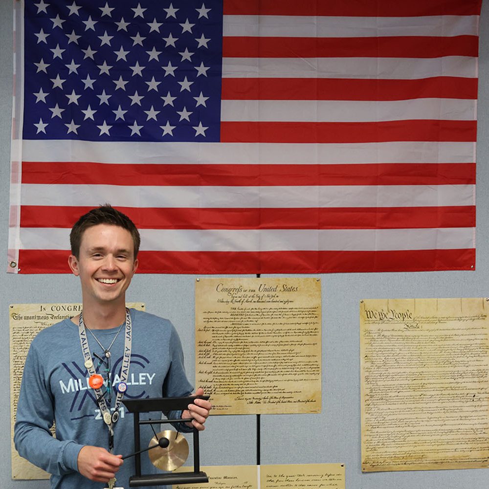 Standing next to the American flag the hangs above his desk, Social Studies teacher Carter Sebasto holds a gong that he was given as a gift. Explaining the value of the gong, Sebasto said, I used to work with a guy who gave me that. He said that I needed it for school to keep the kids in line.