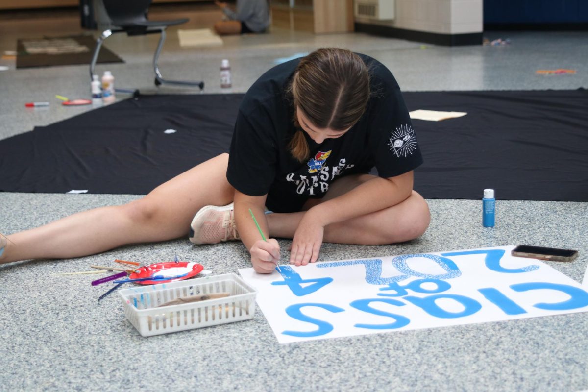 Sitting on the floor, senior Kate Pfeister paints a sign for the senior class hall.