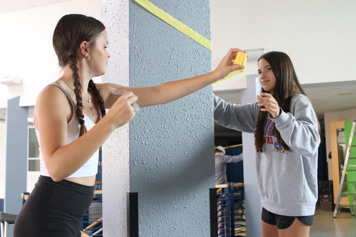 While decorating the halls for spirit week, freshman Ellie Newell and Caitlin Pham pass a paper streamer around the pillar.