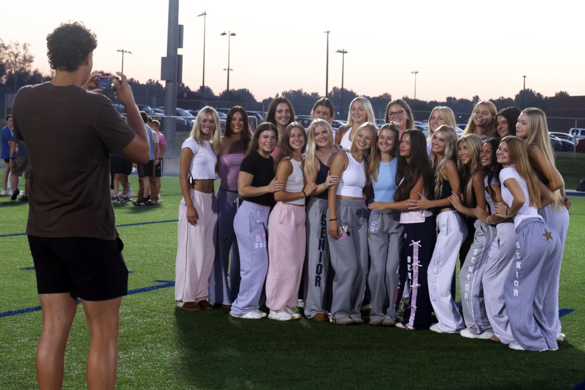 Arms around one another, senior girls friend group smile as senior Carter Crist photographs them.