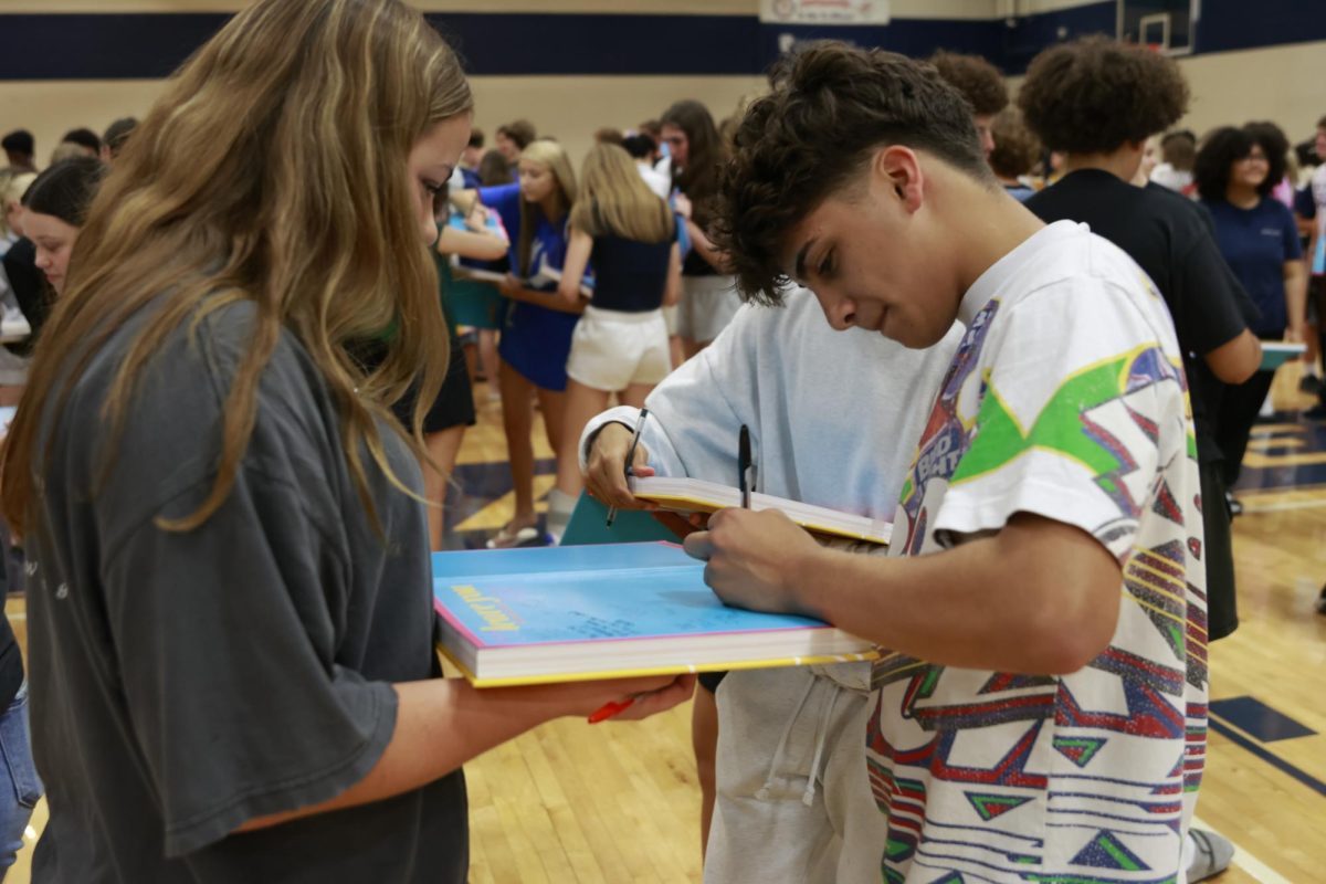 Sophomore Braedyn  Dumlao-Flack holds out her yearbook for sophomore Manny Raso to sign